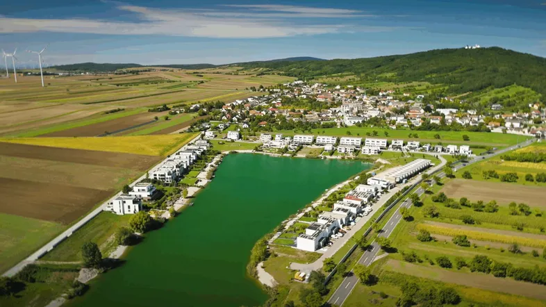 Luftaufnahme einer modernen Wohnsiedlung am Seeufer mit umliegenden Feldern und Hügeln.