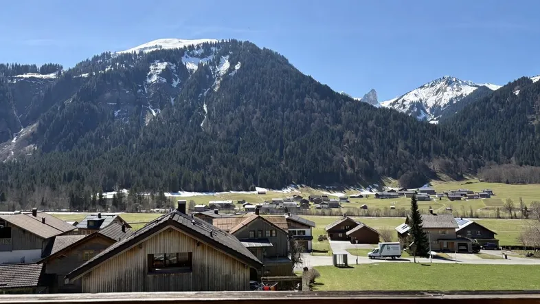 Blick von einem Balkon auf eine malerische Berglandschaft mit bewaldeten Hängen und einem Dorf.