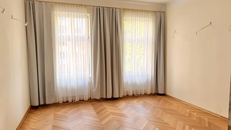 Helles Zimmer mit elegantem Fischgrätparkettboden und zwei großen Fenstern mit Vorhängen.