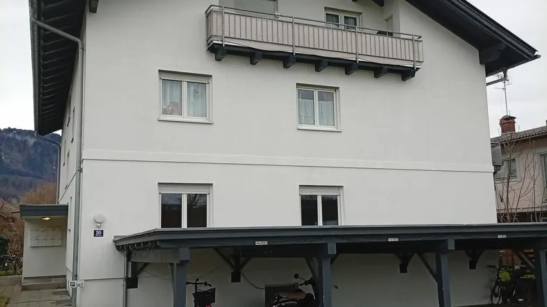 Weißes Mehrfamilienhaus mit dunklem Satteldach, Balkon und überdachtem Carport-Bereich.