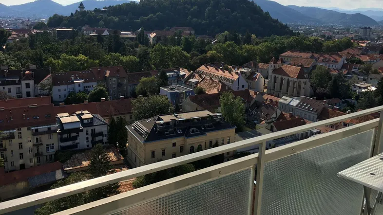 Blick vom Balkon über die Stadt mit grünen Hügeln und Bergen im Hintergrund unter blauem Himmel.