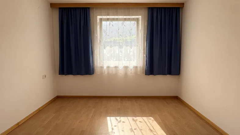 Helles, leeres Zimmer mit Laminatboden und Fenster, das viel Tageslicht hereinlässt.