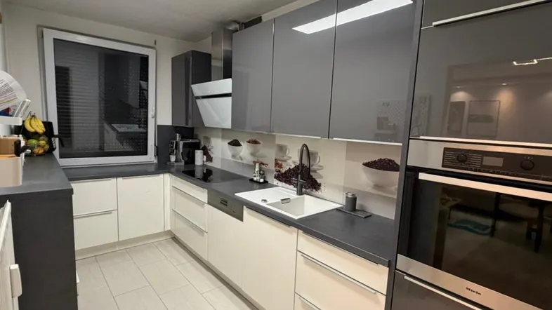 Moderne Küche mit grauen Hochglanzschränken, integriertem Backofen und hellem Fliesenboden.