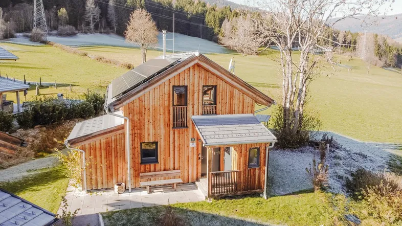 Modernes Holzhaus mit Photovoltaikanlage auf dem Dach, umgeben von grünen Wiesen und Bergen.