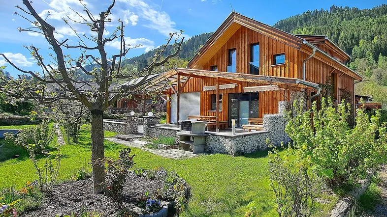 Charmantes Holzhaus mit gepflegtem Garten, Terrasse und Bergblick unter blauem Himmel.