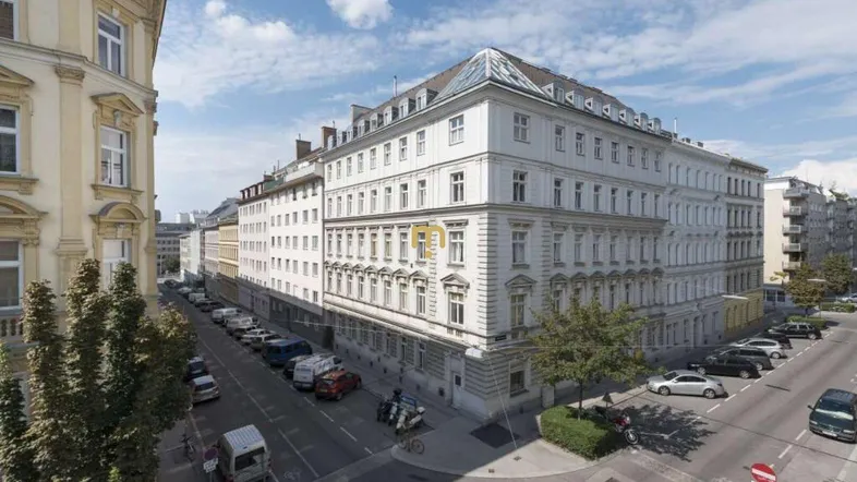Eine Luftaufnahme zeigt ein großes, weißes Eckgebäude mit vielen Fenstern und umliegenden Straßen.