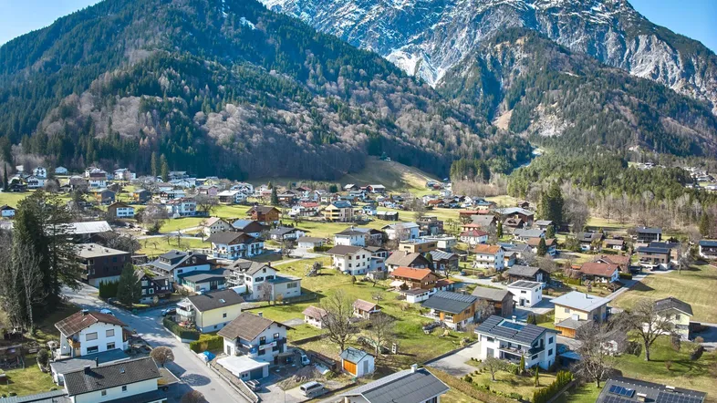 Luftaufnahme eines malerischen Dorfes in einem Tal, umgeben von majestätischen Bergen und grünen Wiesen.