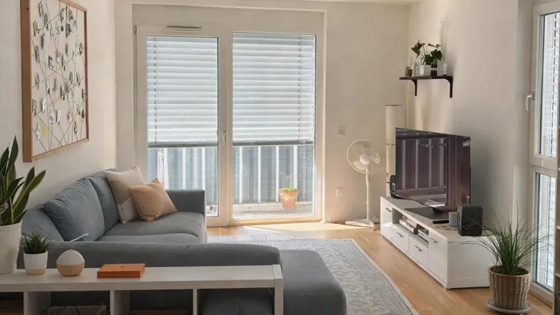 Helles Wohnzimmer mit grauem Ecksofa, Holzboden und Zugang zum Balkon.