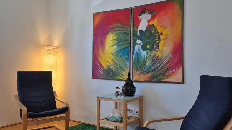 Gemütlicher Wohnbereich mit zwei Sesseln, Holzboden und farbenfrohen Kunstwerken an der Wand, gut beleuchtet.