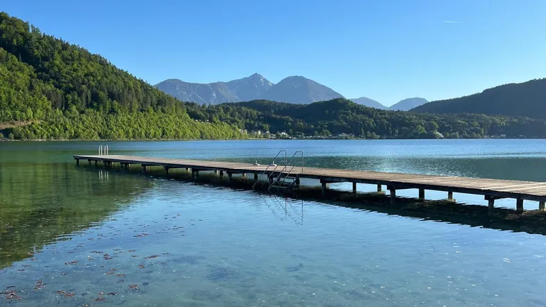 Langer Holzsteg führt in einen klaren See mit bewaldeten Bergen und blauem Himmel im Hintergrund.
