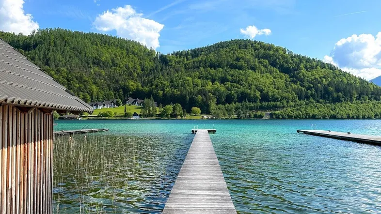 Langer Holzsteg führt in einen klaren, türkisfarbenen See mit grünen Hügeln im Hintergrund.