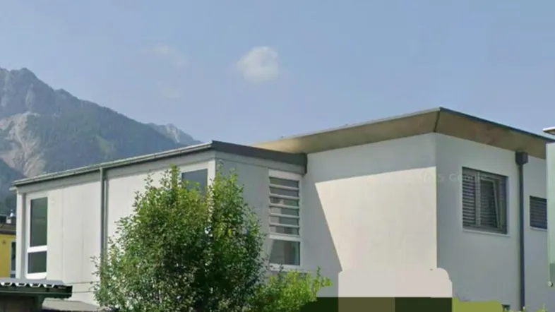 Modernes Gebäude mit Flachdach, umgeben von Bäumen und Bergen im Hintergrund.