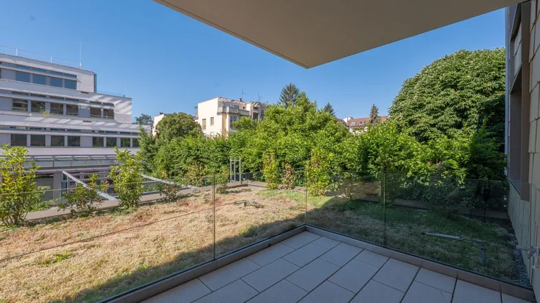 Heller Balkon mit Fliesenboden, Glasgeländer und Blick auf eine begrünte Dachfläche.