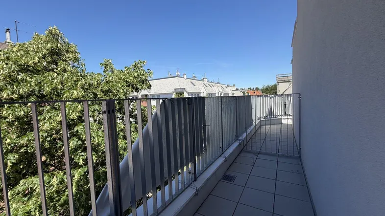Geräumiger Balkon mit Fliesenboden und Blick auf Bäume und umliegende Gebäude unter blauem Himmel.