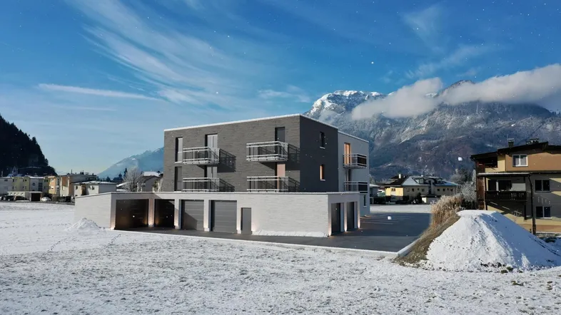 Modernes Mehrfamilienhaus mit Balkonen in winterlicher Landschaft vor einer beeindruckenden Bergkulisse.