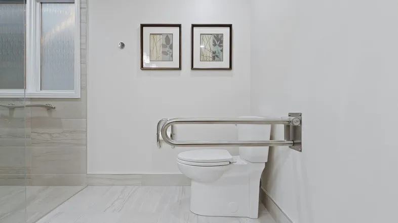 Modernes Badezimmer mit barrierefreier Toilette und Haltegriffen, hell gefliest und mit Dusche.