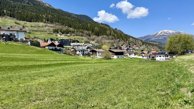 Weitläufige grüne Wiesen mit einem charmanten Dorf im Tal und majestätischen Bergen im Hintergrund unter blauem Himmel.