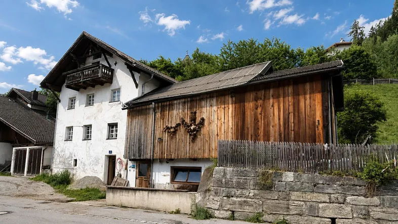 Altes Bauernhaus mit weißer Fassade und rustikalem Holzanbau, umgeben von grüner Natur.