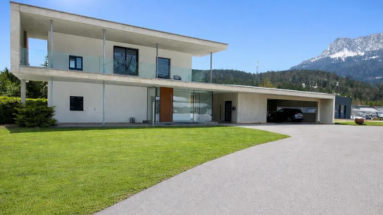 Moderne Villa mit Flachdach, großen Glasflächen, Carport und Bergblick unter strahlend blauem Himmel.