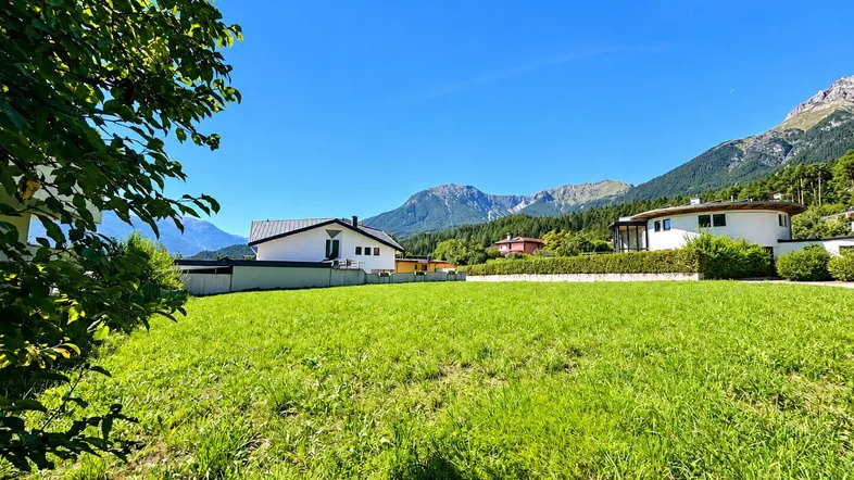 Grüne Wiese mit Bergblick und umliegenden Häusern unter blauem Himmel.