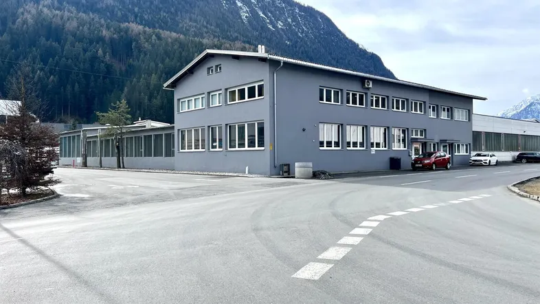 Graues Industriegebäude mit vielen Fenstern vor einer beeindruckenden Bergkulisse und asphaltierter Zufahrt.