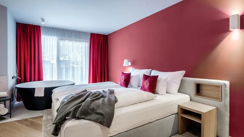 Modernes Schlafzimmer mit freistehender Badewanne, roter Akzentwand und Zugang zum Balkon.