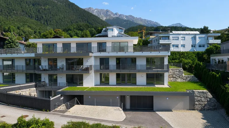Modernes Mehrfamilienhaus mit mehreren Balkonen und Tiefgarage, eingebettet in eine grüne Berglandschaft.