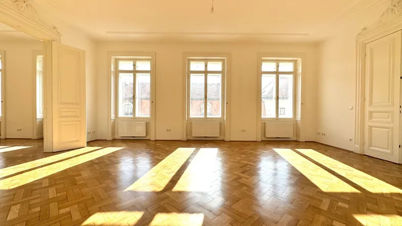 Geräumiges Zimmer mit Fischgrätparkett, hohen Decken und viel natürlichem Licht durch drei große Fenster.