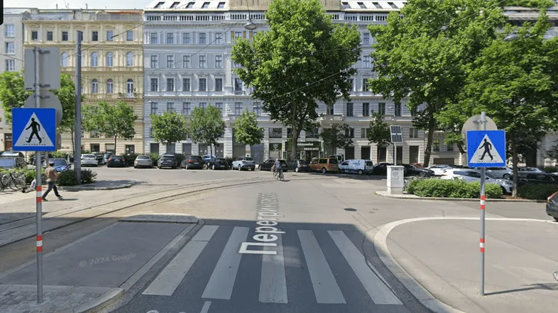 Blick auf die Straße mit historischen Gebäuden, Bäumen und parkenden Autos in einer städtischen Umgebung.