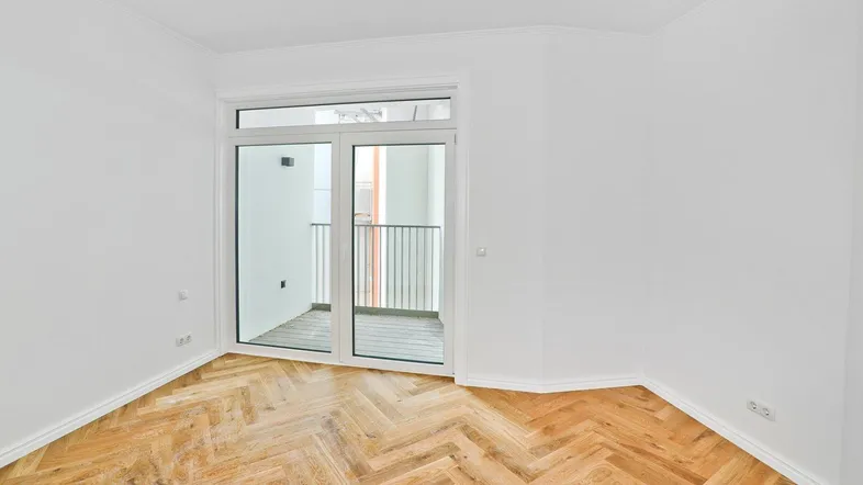 Helles Zimmer mit Fischgrätparkett und Zugang zu einem kleinen Balkon, ideal als Wohn- oder Schlafzimmer.
