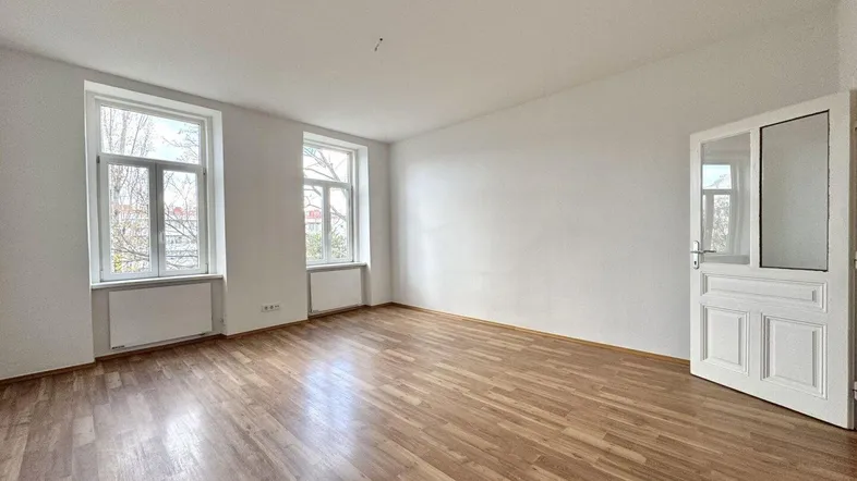 Helles, leeres Zimmer mit Laminatboden und zwei großen Fenstern, die viel Licht hereinlassen.