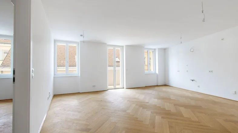 Heller, leerer Raum mit Fischgrätparkettboden und Zugang zu einem französischen Balkon.