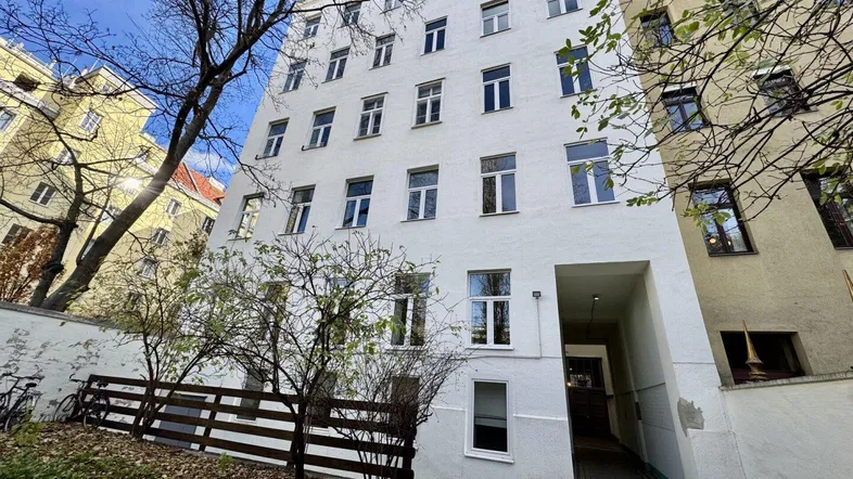 Weißes Mehrfamilienhaus mit Eingangsbereich und Bäumen im Vordergrund.