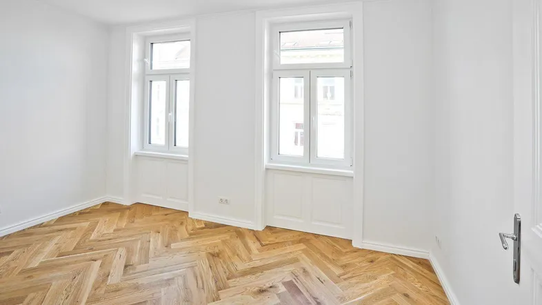 Helles Zimmer mit Fischgrätparkettboden und zwei großen Fenstern, ideal für Wohn- oder Schlafbereich.