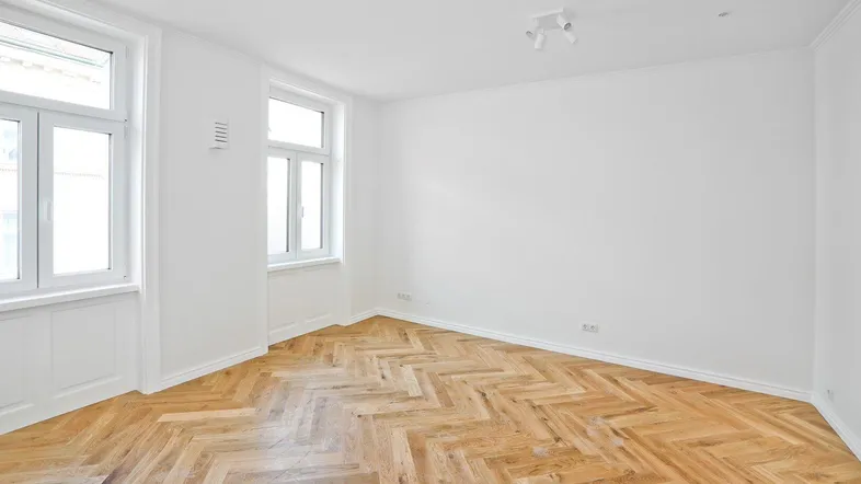 Helles Zimmer mit Fischgrätparkett und zwei großen Fenstern, ideal als Wohn- oder Schlafbereich.