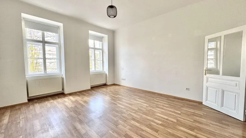 Geräumiges Zimmer mit Laminatboden, zwei Fenstern und einer weißen Tür.