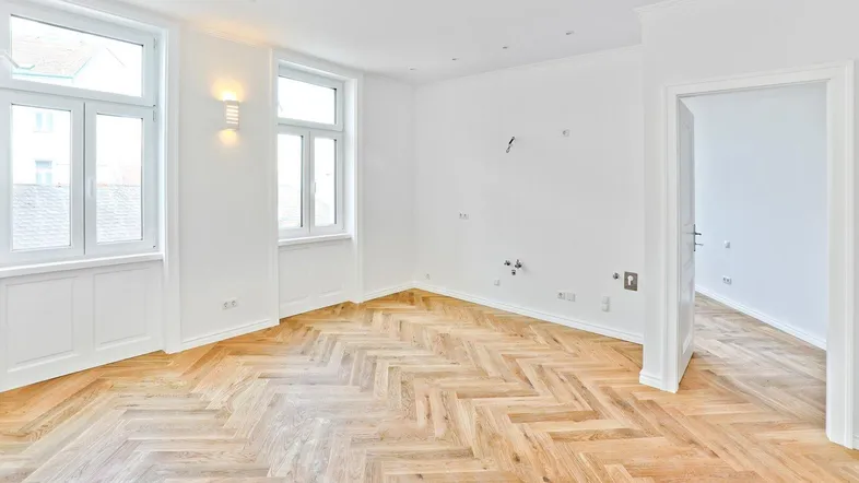 Helles, leeres Zimmer mit Fischgrätparkett und weißen Wänden, bereit zur individuellen Gestaltung.