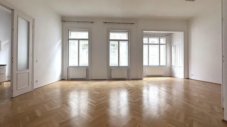 Helles, leeres Zimmer mit Fischgrätparkett und großen Fenstern, die viel Tageslicht hereinlassen.