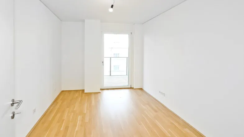 Helles, leeres Zimmer mit Parkettboden und Zugang zu einem kleinen Balkon.