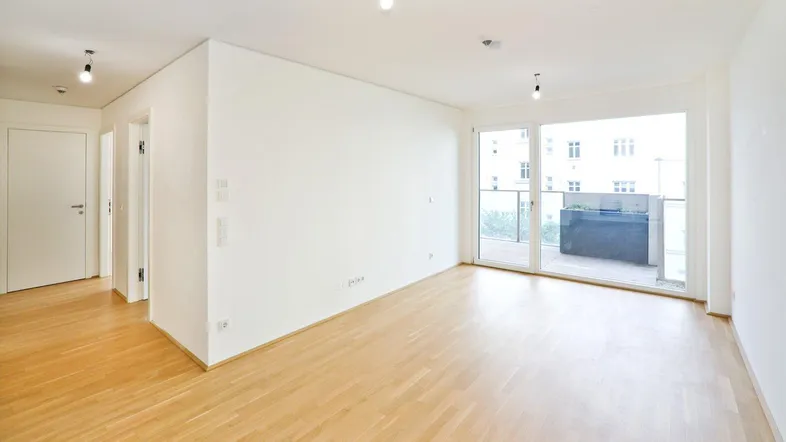 Helles, unfurniertes Wohnzimmer mit Parkettboden und Zugang zu einem Balkon.