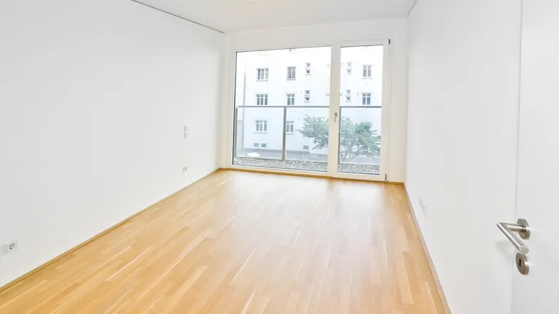 Helles, leeres Zimmer mit Parkettboden und großem Fenster, das Zugang zu einem Balkon bietet.