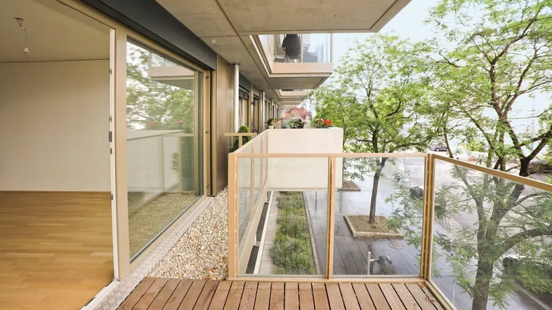 Moderner Balkon mit Holzboden und Glasgeländer, bietet Zugang zu einem hellen Innenraum.