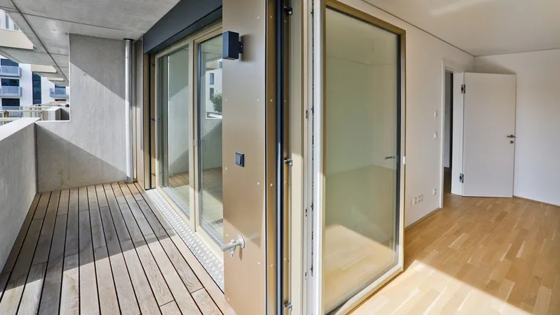 Helles Zimmer mit Holzboden und Zugang zu einem modernen Balkon mit Holzbelag und Glasgeländer.