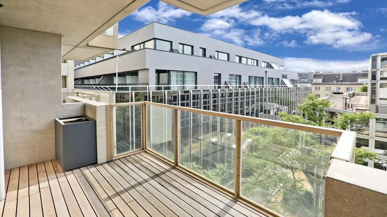 Geräumiger Balkon mit Holzboden und Glasgeländer, bietet einen Blick auf moderne Gebäude und den Himmel.