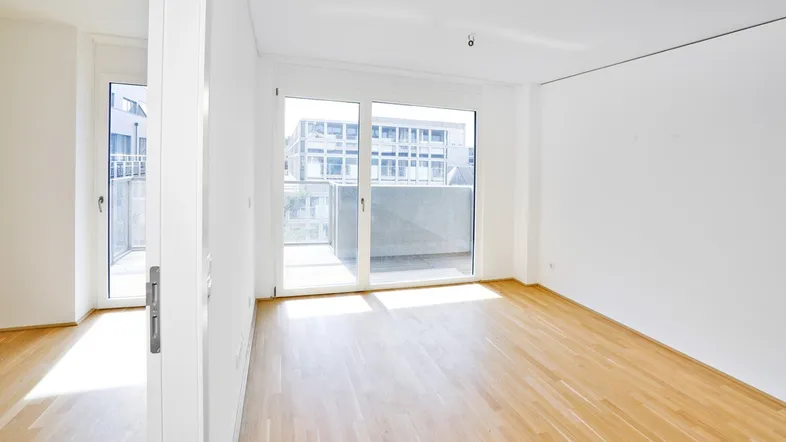 Helles Zimmer mit Holzboden und Zugang zu einem Balkon, ideal als Wohn- oder Schlafbereich.