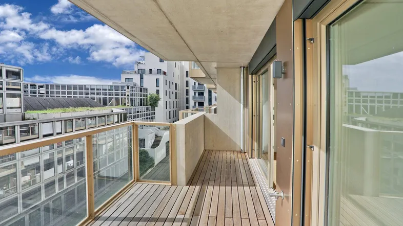 Geräumiger Balkon mit Holzboden und Glasgeländer, bietet einen urbanen Ausblick auf moderne Gebäude.