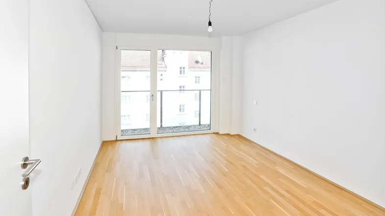 Helles, leeres Zimmer mit Parkettboden und Zugang zu einem kleinen Balkon.