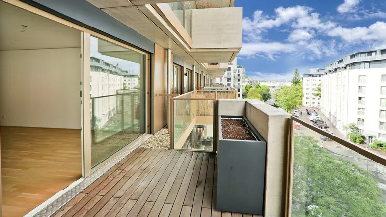 Geräumiger Balkon mit Holzfußboden, Glasgeländer und Blick auf die Stadt und umliegende Gebäude.