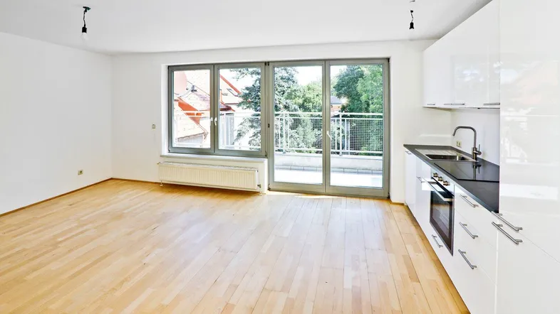 Heller offener Wohnbereich mit integrierter Küche, Parkettboden und Zugang zum Balkon.