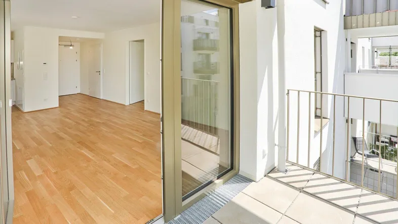 Heller Innenraum mit Holzboden und Zugang zu einem sonnigen Balkon, der einen Blick auf die Umgebung bietet.